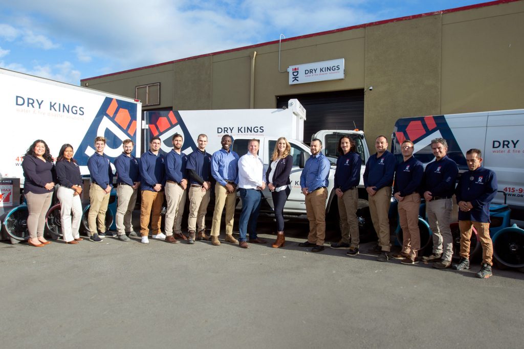 A diverse group of sixteen employees stands in front of "Toronto Restoration" business with vans and flags, smiling, dressed in coordinated uniforms under a clear sky.