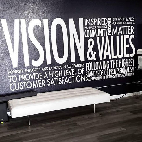 A modern office in Toronto with a dark wall featuring large white text that reads "vision & values," accompanied by various inspirational phrases. A sleek white bench sits below the text.