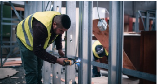 A construction worker in a reflective vest uses a power drill on metal framing at a Toronto restoration site, with another worker in the background.