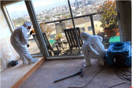 Two workers in protective suits and masks clean a room with exposed walls and construction tools, with a cityscape visible through an open balcony door during a Toronto restoration.