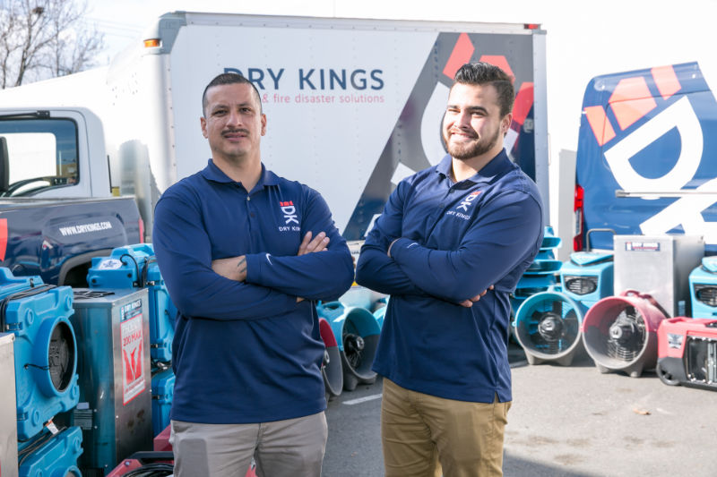 Two men in work uniforms standing confidently with crossed arms in front of a service truck and equipment labeled "Toronto Restoration," under a clear sky.