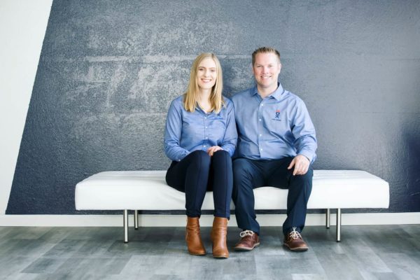 A man and a woman, both in blue shirts, smiling and seated together on a white bench against a textured gray wall in Toronto.