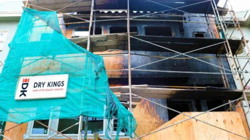 A damaged building under restoration in Toronto, covered with green netting and scaffolding, showing signs of fire damage with charred exteriors. A sign reading "Dry Kings" is visible on the scaff