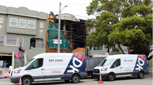 Two dry kings company vans parked in front of a building under renovation covered with scaffolding, in a Toronto street lined with trees.