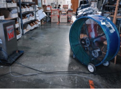 Large blue industrial fan on wheels positioned on a concrete floor in a Toronto restoration warehouse, surrounded by boxes and shelving units filled with supplies.