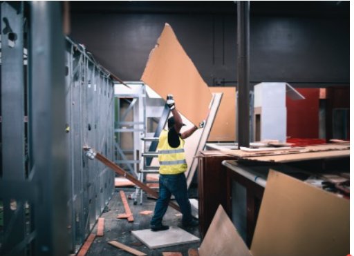 A construction worker in a yellow vest and helmet carries a large piece of drywall inside a building under renovation with visible steel studs and scattered debris in Toronto.